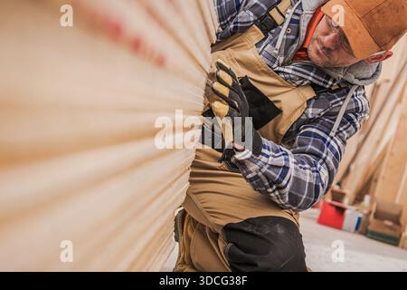 Man focuses on sanding wood while kneeling in a workshop environment with wooden panels around. Stock Photo