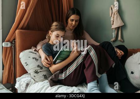 Mother sitting with daughter reading book on bed at home Stock Photo