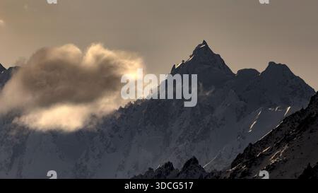 View of snow-capped peaks piercing through the misty veil, sun rays filtering through clouds in Hunza, Gilgit Baltistan, Pakistan. Stock Photo