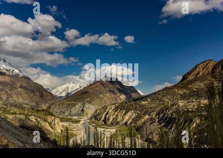 View of rugged, sunlit mountains casting shadows on the valley, while snow-capped peaks glisten under a bright blue sky dotted with fluffy clouds, Hun Stock Photo