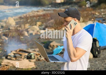 Profile view of a man in cap on a phone call, clutching an open laptop near tent, campfire, and rocky river edge. Stock Photo