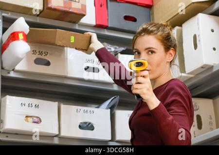 A woman in maroon shirt pulls a brown cardboard box from shelf while aiming a yellow-tipped barcode scanner forward in storage area. Stock Photo
