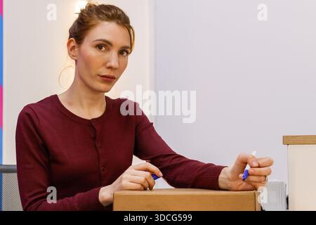 Side view of female worker gripping blue pen directed towards a brown carton on the counter. Stock Photo