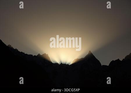 View of the sun's radiant fingers reaching out from behind the dark, jagged mountain peaks in the golden hour, Passu, Gilgit Baltistan, Pakistan. Stock Photo