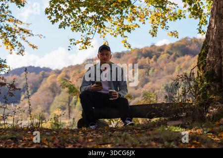 Individual in light jacket sits on log bench with coffee in hand against backdrop of autumn trees and distant hills. Stock Photo