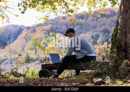 Side view of man in cap sitting on log, hand on coffee cup, typing on open laptop, autumn trees and hills in background Stock Photo