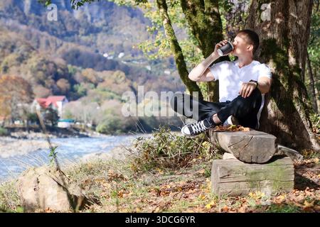 Man in white shirt and black pants sits cross-legged on wooden log, drinks from black mug, with autumn forest, river, and distant buildings in backgro Stock Photo