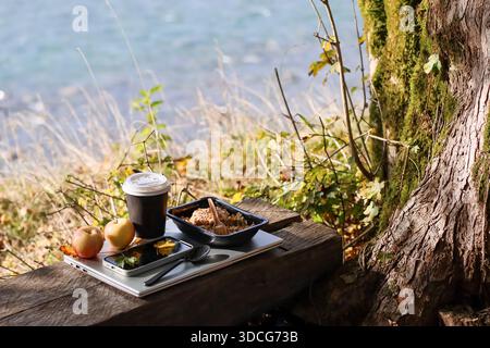 Two apples, dark coffee cup with white lid, black container with grain mix and fork, small snack trays with spoon on silver laptop placed on wooden be Stock Photo