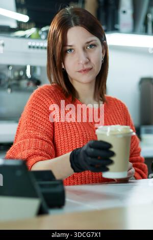 Smiling woman in orange sweater and black gloves offers foamy beverage in disposable cup at cafe counter. Stock Photo
