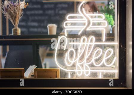 Bright neon coffee sign with cup and steam on cafe window cozy interior and dried flowers in background Stock Photo