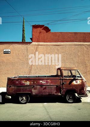Old rusty pickup truck parked on the street in front of a textured beige wall under a clear blue sky with electric wires overhead Stock Photo