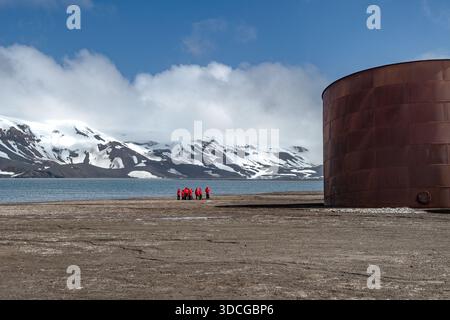 View of a group of people in red jackets stand small against the stark landscape, with the cold blue water and snow-capped mountains beyond, Deception Stock Photo