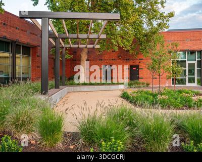 View of a tranquil courtyard where a modern pergola casts shadows on a brick wall amidst lush greenery, Apollo Park, Oregon, United States. Stock Photo