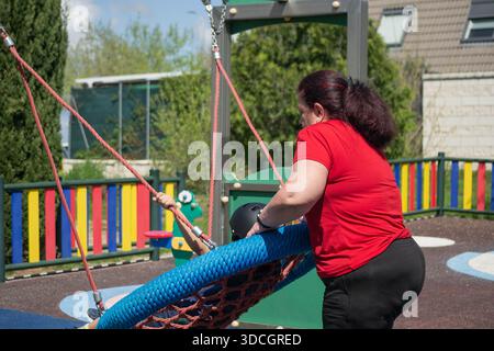 Woman assisting child wearing protective helmet while using swing at playground Stock Photo