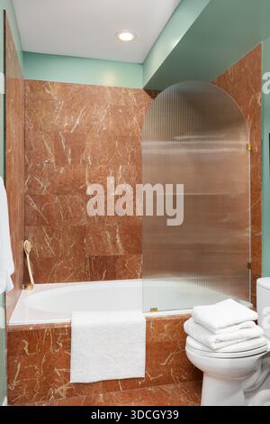 A luxurious bathroom featuring red marble tiled walls, white bathtub with a ribbed glass shower screen, and teal paint. Stock Photo
