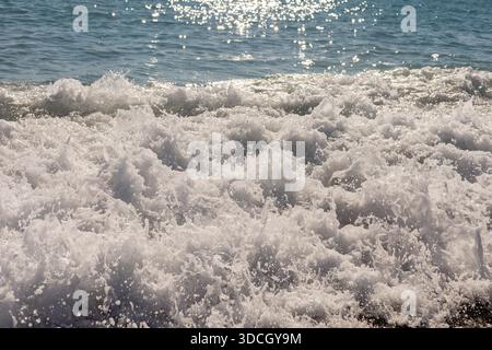 Bright sunlight reflects on the ocean surface as waves crash on the shore. Water splashes create foam and movement. The scene captures a lively moment Stock Photo