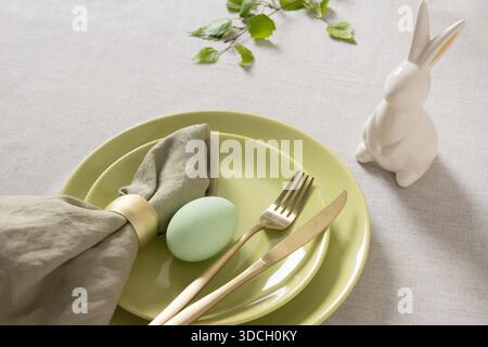 Minimal Easter place setting with pastel egg, green ceramic plates, golden cutlery and white bunny figurine on neutral tablecloth, spring holiday concept. Stock Photo