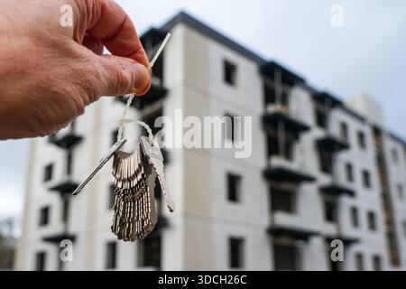 Hand holding a set of keys in front of modern apartment building under construction, symbolizing property handover, housing development and investment Stock Photo
