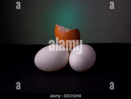 Three eggs arranged with a broken brown eggshell on top, set against a dark background. Stock Photo