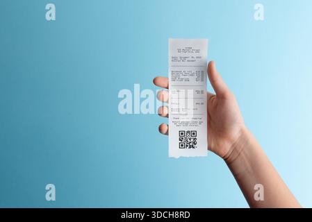 Hand holding paper receipt with qr code on blue background, representing digital payments, expense documentation, receipt scanning, and paperless acco Stock Photo