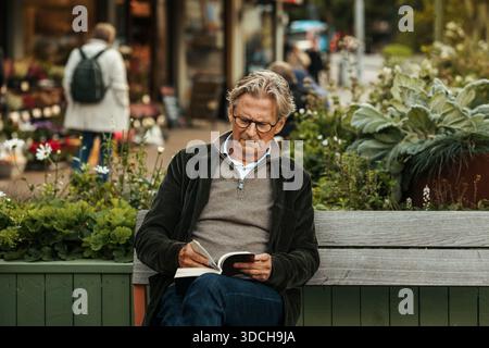 Senior man reading book while sitting on bench at street Stock Photo
