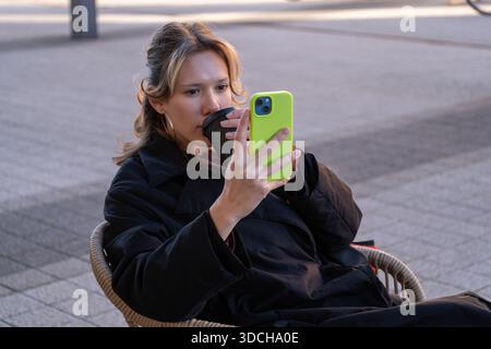 A young female student sitting outdoors and drinking takeaway coffee while using a smartphone. This image is suitable for student lifestyle concepts, Stock Photo