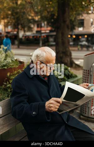 High angle view of concentrated senior man reading newspaper while sitting on bench at street Stock Photo