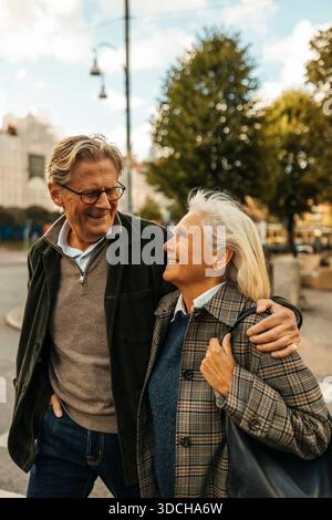 Happy elderly man walking with arm around woman while strolling at city street Stock Photo