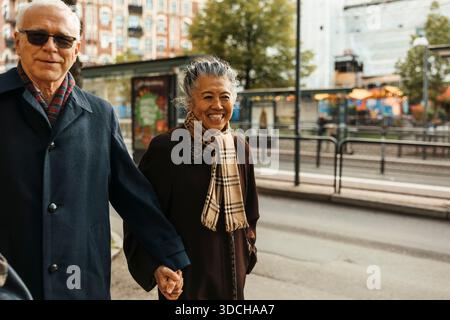 Smiling senior woman holding hands of man while walking on sidewalk in city Stock Photo