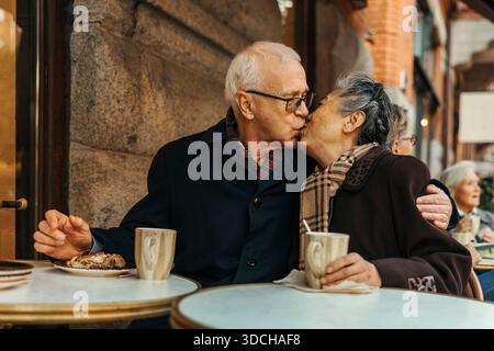 Senior couple kissing each other while sitting at table in sidewalk cafe Stock Photo