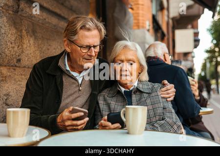 Elderly couple using smart phone while sitting at table in sidewalk cafe Stock Photo
