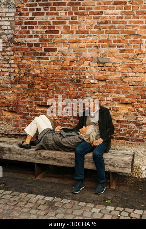 Carefree senior woman lying down on man's lap at bench near brick wall Stock Photo