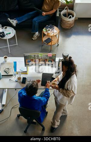 High angle view of female tech expert talking with male colleague working on desktop PC in startup office Stock Photo