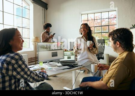 Happy female programmer talking with colleagues while sitting at desk in tech hub Stock Photo