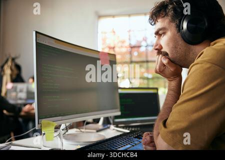 Thoughtful male programmer wearing wireless headphones looking at codes on computer in startup office Stock Photo