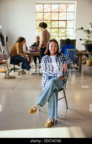 Portrait of confident female tech entrepreneur sitting on chair with legs crossed at knee in startup office Stock Photo