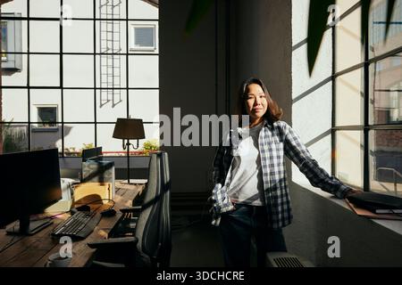 Portrait of confident young female entrepreneur standing with hand in pocket near window at tech office Stock Photo