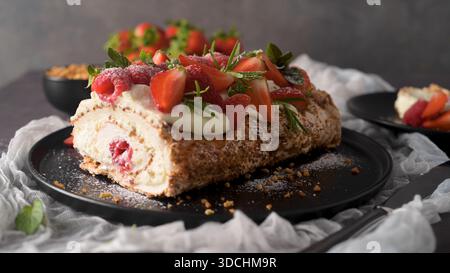 A delicate meringue roll filled with rich cream and fresh raspberries is displayed on a black plate. The dessert is topped with sliced strawberries, m Stock Photo