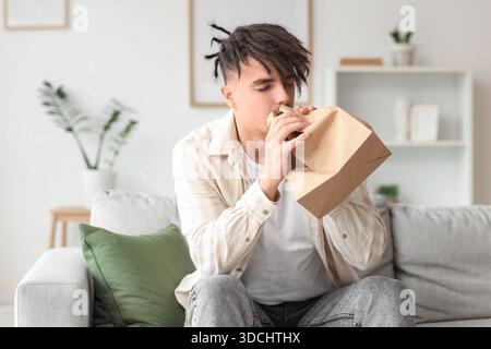 Young man with paper bag having panic attack on sofa at home Stock Photo