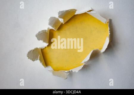 A piece of white paper with a jagged circular hole torn through it, revealing a yellow color background. The curled edges and contrast create a 3D vis Stock Photo
