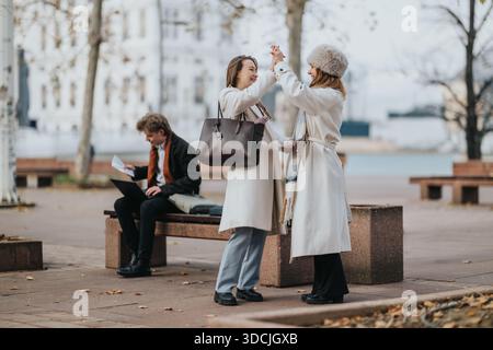 Two women in winter coats high-five in a city park among friends on a cold day Stock Photo