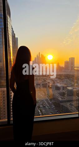 Woman Admiring Dubai Skyline at Sunset. A silhouetted woman stands by a high-rise window overlooking the glowing Dubai skyline during a peaceful Stock Photo