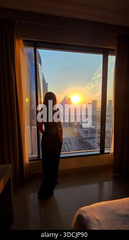 Woman Watching Dubai Sunset from Hotel Window. Silhouette of a woman gazing out a high-rise hotel window at the Dubai skyline during a vibrant urban Stock Photo