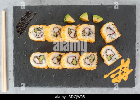 Two rows of deep-fried sushi rolls with crab and cucumber garnished with sauces and black sesame seeds. Stock Photo