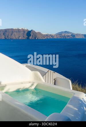 View of a pristine turquoise pool contrasting with the stark white architecture against the deep blue Aegean Sea and distant rocky cliffs, Oia, Thira, Stock Photo
