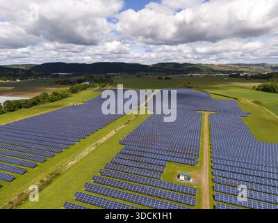 Aerial view of expansive solar panels reflecting the sky, juxtaposed against the bright green fields and distant hills under a cloudy sky, Perth, Scot Stock Photo