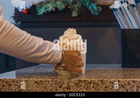 Horizontal medium shot eye level view of a woman's hand with a charm bracelet holding a large white mug topped with whipped cream in a coffee shop Stock Photo