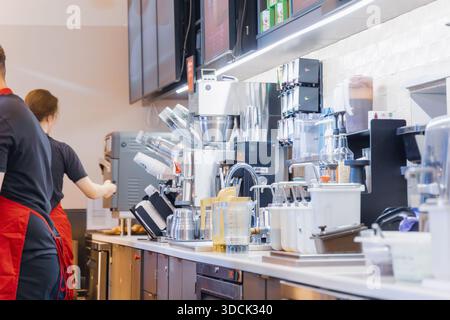 Two cafe workers in uniform preparing drinks at a busy commercial coffee station with espresso machines and equipment Stock Photo