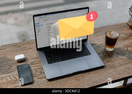 A modern workspace with a laptop displaying an email inbox and a new message notification, smartphone, wireless earbuds, and a glass of coffee on a wo Stock Photo