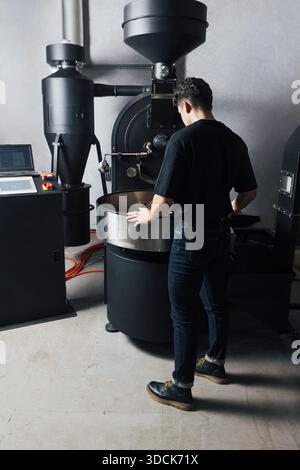 Full length of a male worker at an industrial roasting coffee machine. Young man controlling the roasting coffee process. Stock Photo